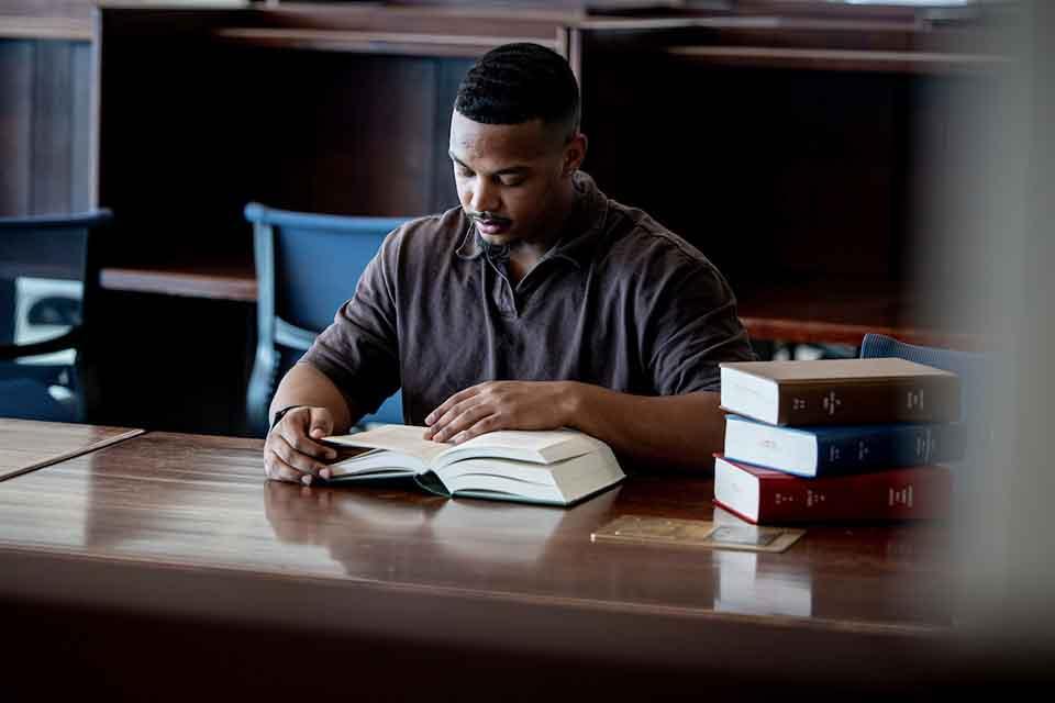 A male student reads from a book at a table in thel library.